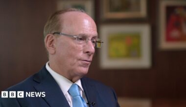 Wide shot showing Blackrock CEO Larry Fink being interviewed by BBC business editor Simon Jack. The two men are sitting across from each other with a long wooden table behind them and four pieces of art hanging on the wall