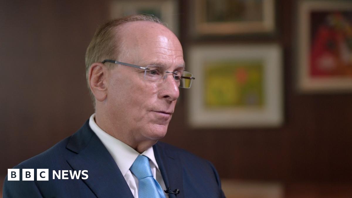 Wide shot showing Blackrock CEO Larry Fink being interviewed by BBC business editor Simon Jack. The two men are sitting across from each other with a long wooden table behind them and four pieces of art hanging on the wall