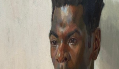 A head and shoulders portrait of the artist Nahem Shoa shows him wearing a plain red top, looking slightly off to the right, his gaze fixed on somthing or somone we can't see.