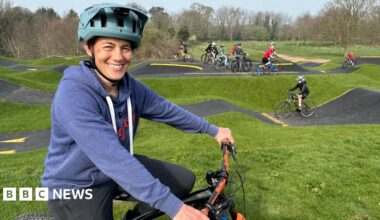 A woman wearing a mid-blue hoodie and a turquoise cycle helmet sits on a bike and smiles at the camera. In the background a number of children are riding bikes on a pump track, a bike track with lots of hills of various sizes.