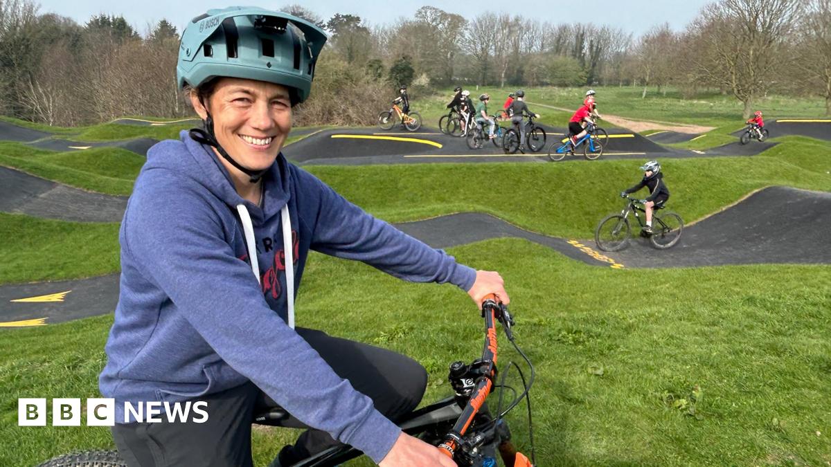 A woman wearing a mid-blue hoodie and a turquoise cycle helmet sits on a bike and smiles at the camera. In the background a number of children are riding bikes on a pump track, a bike track with lots of hills of various sizes.