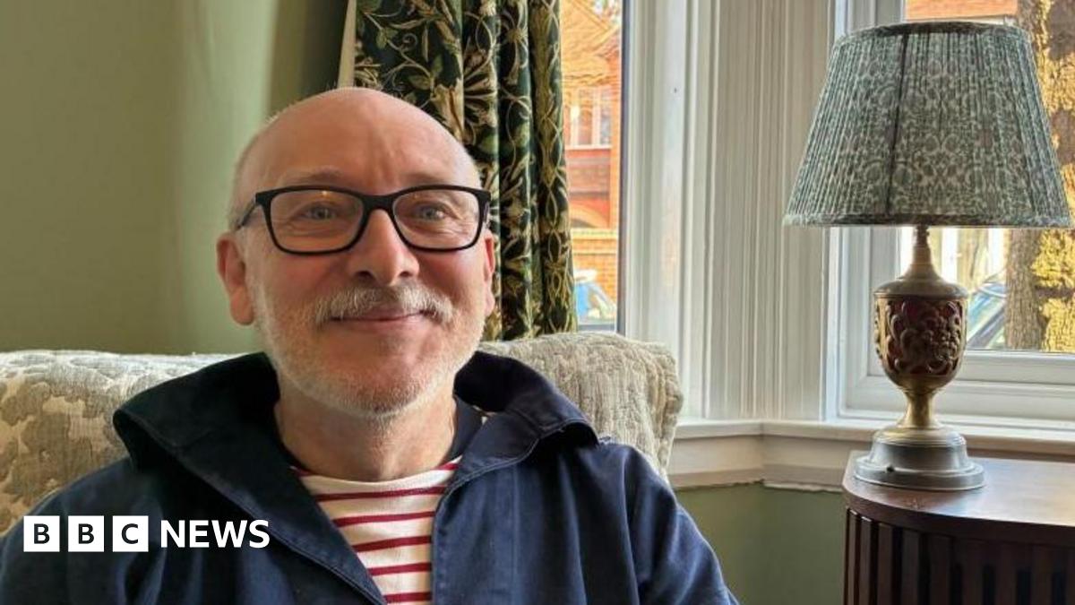 Andy Hughes sitting in front of his front window at home in Nuneaton. A lamp sits on the table behind him and neighbouring homes and parked cars can be seen outside the window in the morning sunshine.