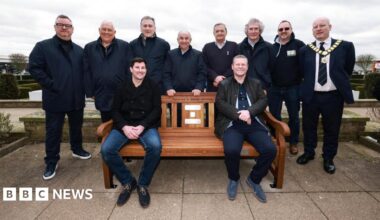 Several people gather around a brown wooden bench. Eight people are stood behind it, with two people sat on the bench. On the bench there are several plaques, and above them is some black writing which says "someone is always listening".