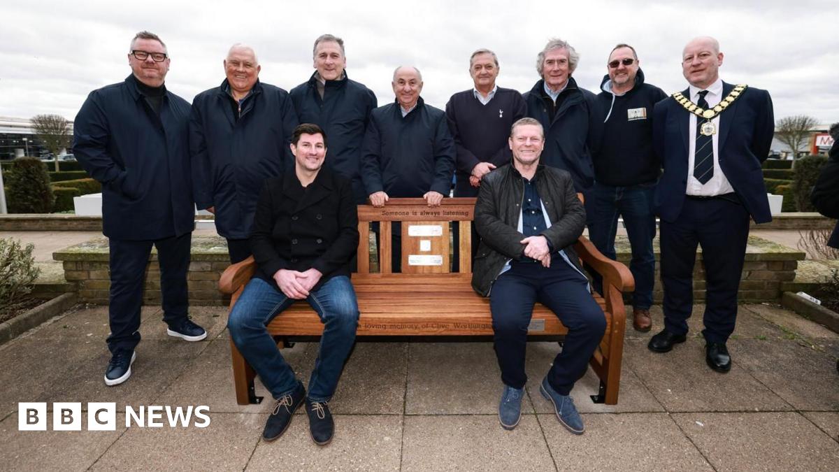 Several people gather around a brown wooden bench. Eight people are stood behind it, with two people sat on the bench. On the bench there are several plaques, and above them is some black writing which says "someone is always listening".
