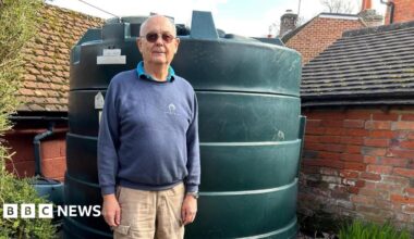 Chris Wheeler stands next to his dark green heating oil tank in his garden. He is wearing glasses, a blue sweatshirt and beige cargo trousers. The cylinder-shaped tank is in the corner of the garden in front of an old brick wall.