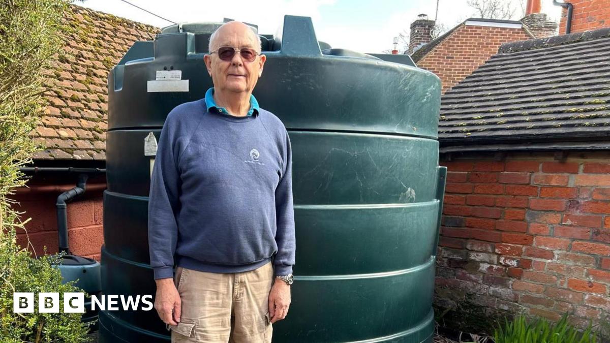 Chris Wheeler stands next to his dark green heating oil tank in his garden. He is wearing glasses, a blue sweatshirt and beige cargo trousers. The cylinder-shaped tank is in the corner of the garden in front of an old brick wall.