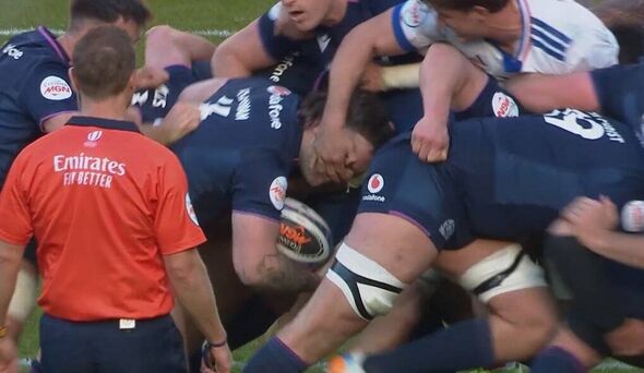 A group of rugby players, wearing blue and white uniforms, are engaged in a scrum on the field. An official, dressed in an orang