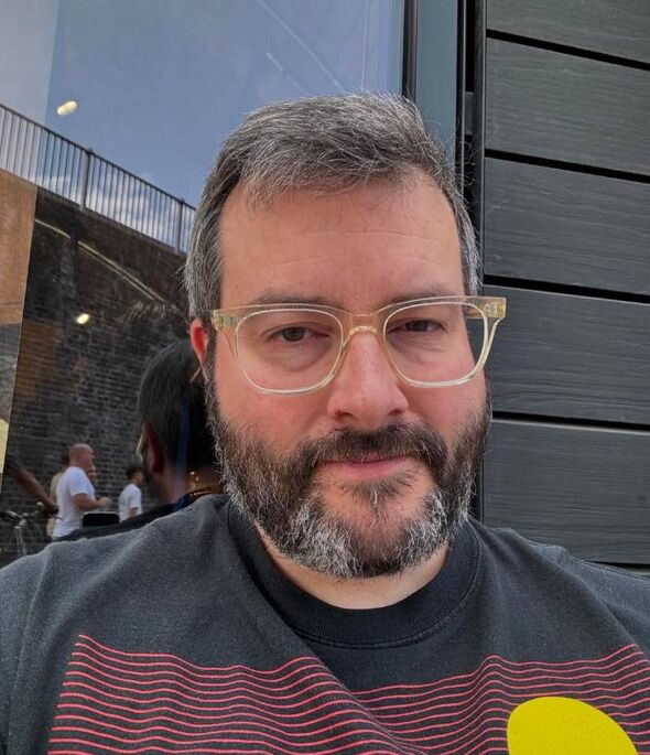 An individual with a beard and mustache, donning a black shirt and clear-framed glasses, stands in front of a textured, dark-col An individual with a beard and mustache, donning a black shirt and clear-framed glasses, stands in front of a textured, dark-col