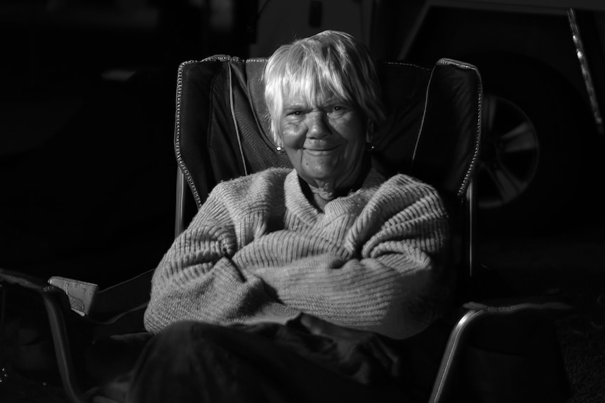 A black-and-white photo of an elderly woman sitting on a camp chair at night smiling at the camera.