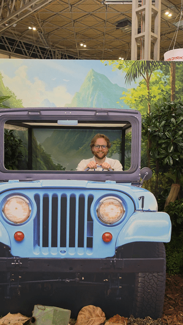 Man smiling behind the wheel of a light blue vintage-style jeep display, surrounded by tropical plants with a painted jungle and mountain backdrop inside an exhibition hall.