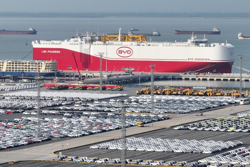 Rows of vehicles lined up at a port near a large white and red ship with a logo saying "BYD".