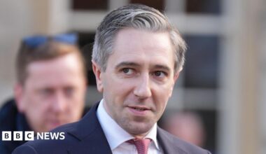 A close up shot of a man with grey hair, wearing a suit standing outside. Another man can be seen out of focus in the background.