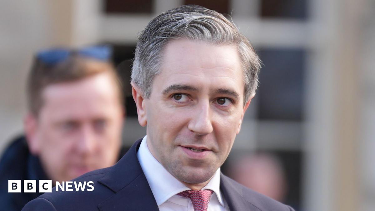 A close up shot of a man with grey hair, wearing a suit standing outside. Another man can be seen out of focus in the background.
