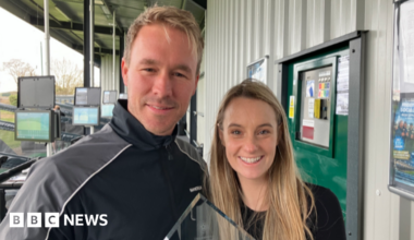 A man and woman, both with blonde hair, smile at the camera. They are holding a glass award between them.  Behind them are bays of a golf driving range