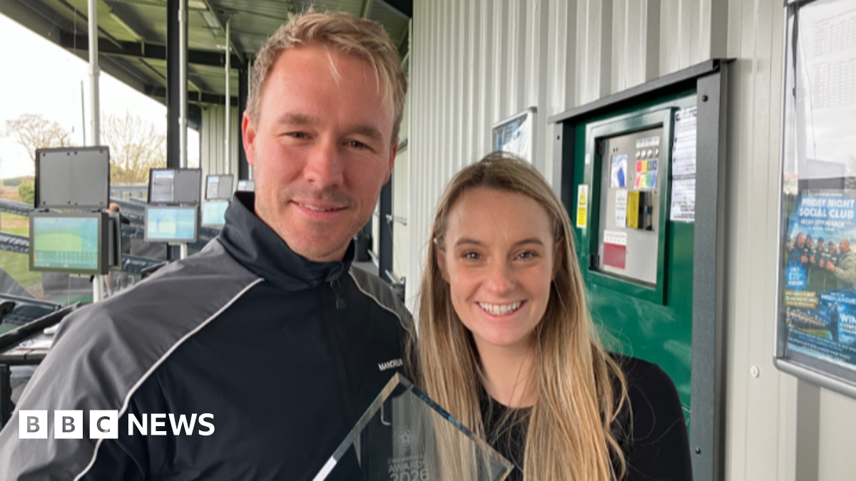 A man and woman, both with blonde hair, smile at the camera. They are holding a glass award between them.  Behind them are bays of a golf driving range