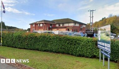 A view from the road looking at the main building on the golf course which has a large hedge in front of it. The building itself is made up of red brick and is two storeys high. There are a number of vehicles parked in front of the property.