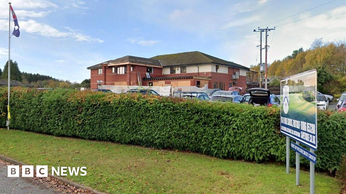 A view from the road looking at the main building on the golf course which has a large hedge in front of it. The building itself is made up of red brick and is two storeys high. There are a number of vehicles parked in front of the property.