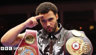 A side-on image shows boxers Moses Itauma and Dillian Whyte from waist up during a fight in August 2025. Itauma is on the right of the image landing a right-handed jab through the middle of Whyte's defences onto his chin, leading his opponent to turn and duck his head towards the camera and screw up his face. The bearded Itauma is wearing black shorts and has black gloves with a red wrist strap, whereas Whyte wears black and purple gloves and shorts in a similar design which also have his surname on the waistband