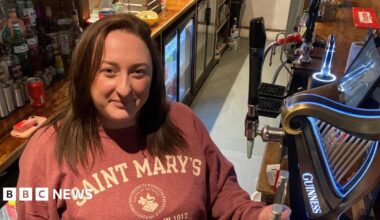Megan stands behind the bar, smiling up at the camera. Behind her are bottles of alcohol and she stands next to the handles to pull pints. She has shoulder length brown hair and wears a salmon coloured hoodie with white writing on.