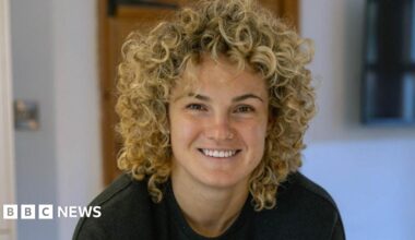 A woman with curly blonde hair smiles at the camera. She is wearing a black T-shirt.