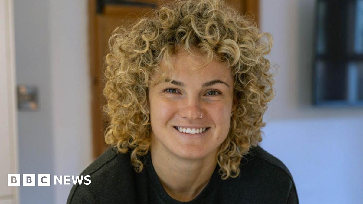 A woman with curly blonde hair smiles at the camera. She is wearing a black T-shirt.