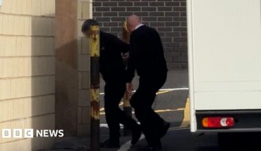 A man being led away from a van by another man wearing a black jumper and a shirt. He is walking into a brick building.