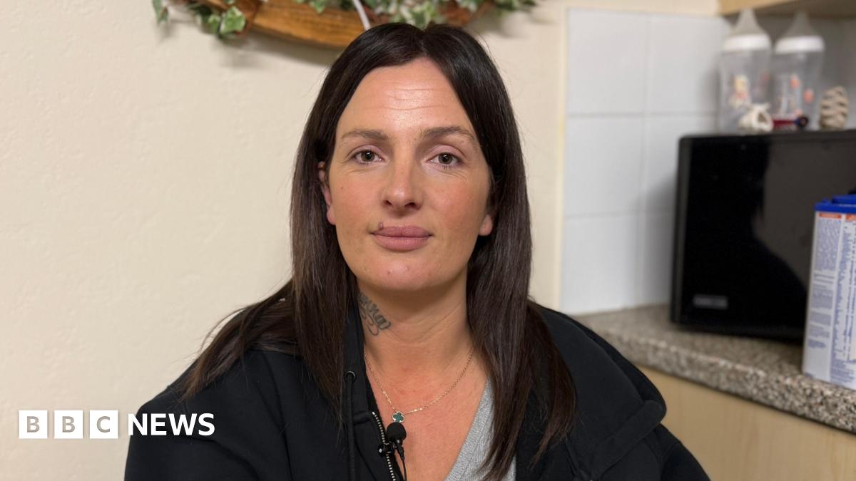 Krystal Ogier, a middle-aged woman, wearing a neutral expression. She is wearing a black hoodie with a grey T-shirt underneath. She has a tattoo on her neck and long, dark brown hair.