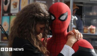 Tom Holland as Spider-Man holds Zendaya, who plays MJ, on set. Holland is dressed in a ful Spider-Man suit against a New York backdrop and Zendaya wears a black top and has long brown hair