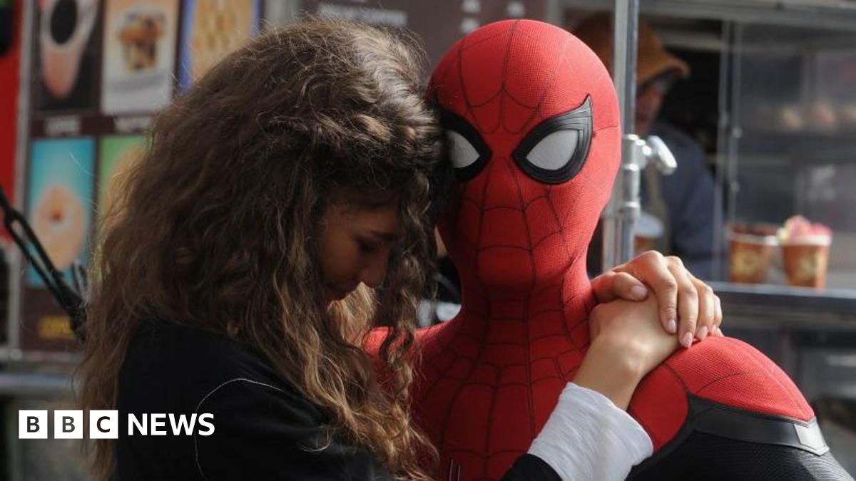 Tom Holland as Spider-Man holds Zendaya, who plays MJ, on set. Holland is dressed in a ful Spider-Man suit against a New York backdrop and Zendaya wears a black top and has long brown hair