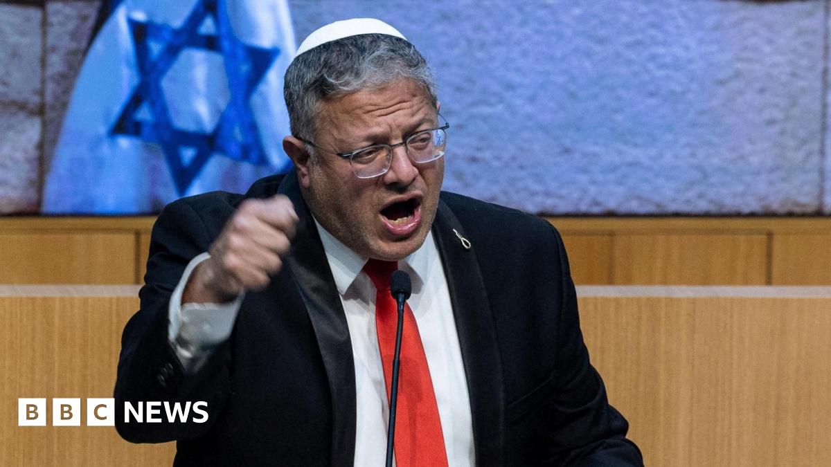 Itamar Ben-Gvir wearing a black suit, red tie and white kippah, talking on a podium in front of Israeli flags