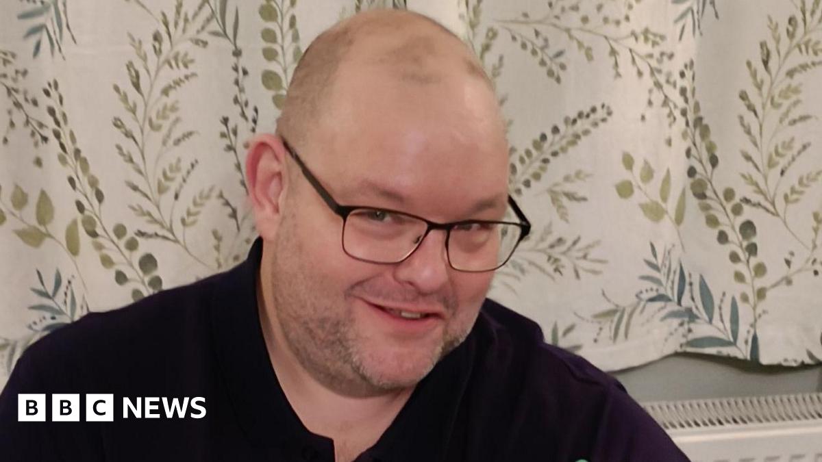 A man, Gary Stanley, smiles into the camera. He is wearing black glasses and a black top. Behind him are white curtains with details of green leaves and branches.