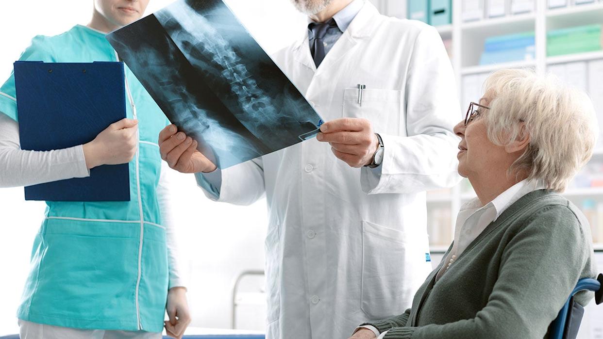 Older woman sitting in wheelchair as doctor and nurse analyze her x-ray results showing spine.