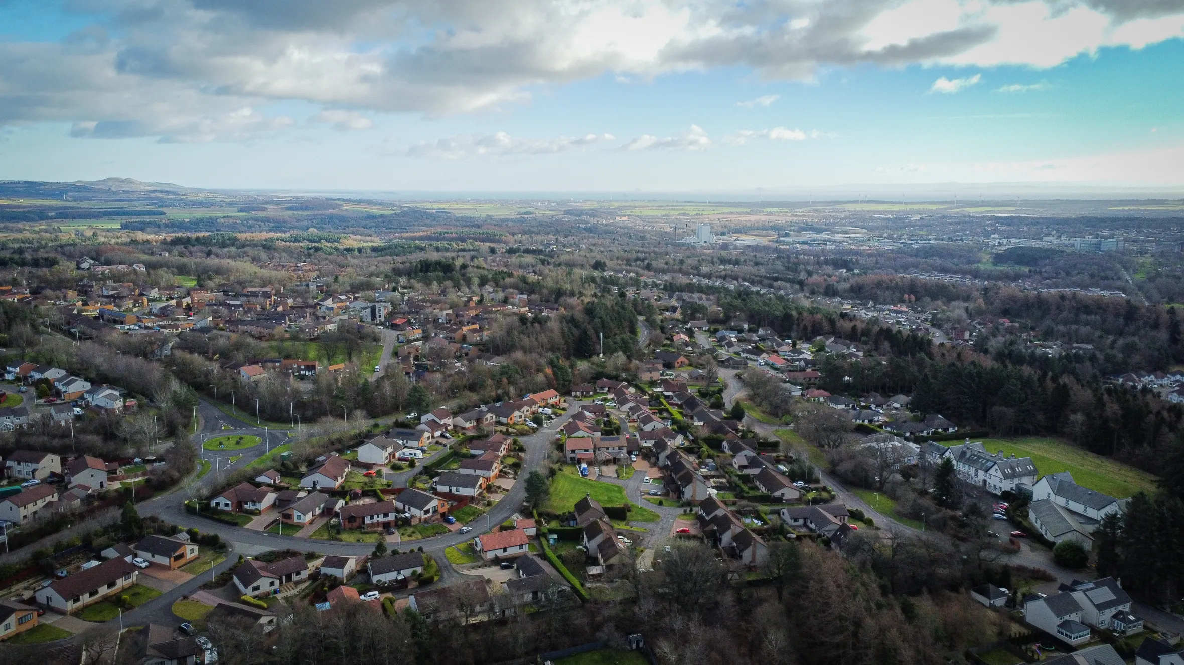 Aerial view of North Glenrothes, showing houses surrounded by trees and a circular road, with distant hills and a body of water under a cloudy sky.