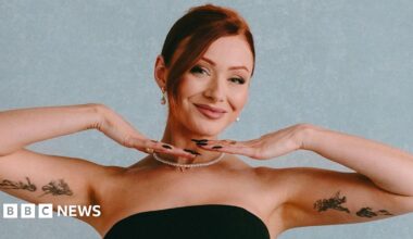 Elz the Witch poses in front of a light blue background. She's wearing a strapless black gown and a thing string of pearls. She's smiling, her hands under her chin, framing her face, and showing off the tattoos of figures drawn in intricate black ink lines on the inside of her upper arms.