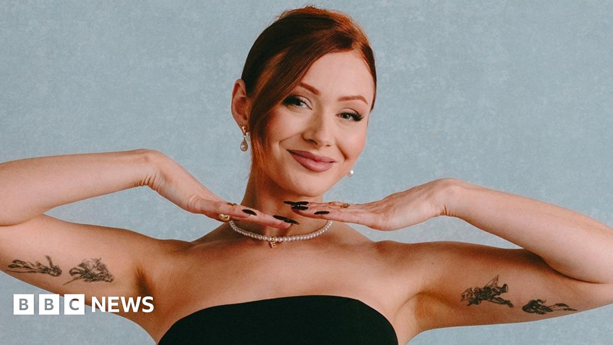 Elz the Witch poses in front of a light blue background. She's wearing a strapless black gown and a thing string of pearls. She's smiling, her hands under her chin, framing her face, and showing off the tattoos of figures drawn in intricate black ink lines on the inside of her upper arms.