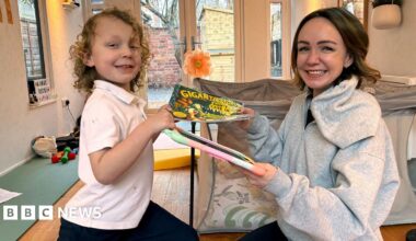 Romi, who has blond curly hair and is wearning a white t-shirt, is passing a book to his mum, Alexis who has short brown hair and is wearning a grey hoodie. She is passing a digital device with a colourful cover back to him and both are smiling.