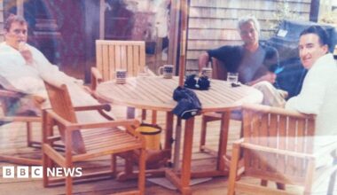 A woman with dark hair and a multiple colour jumper siting on a green sofa inside her living room.
