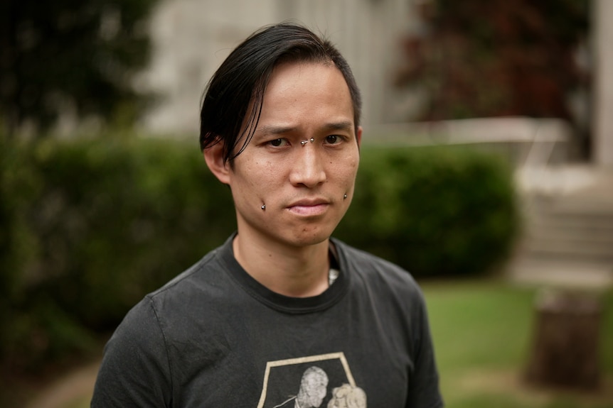 William, a young man with piercings in each cheek, above the nose, looks seriously into the camera.