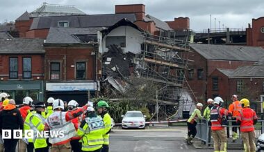 A partially collapsed building that shows roof tiles strewn over a pile of debris a scaffold hangs nearby. A number of emergency workers stand nearby.