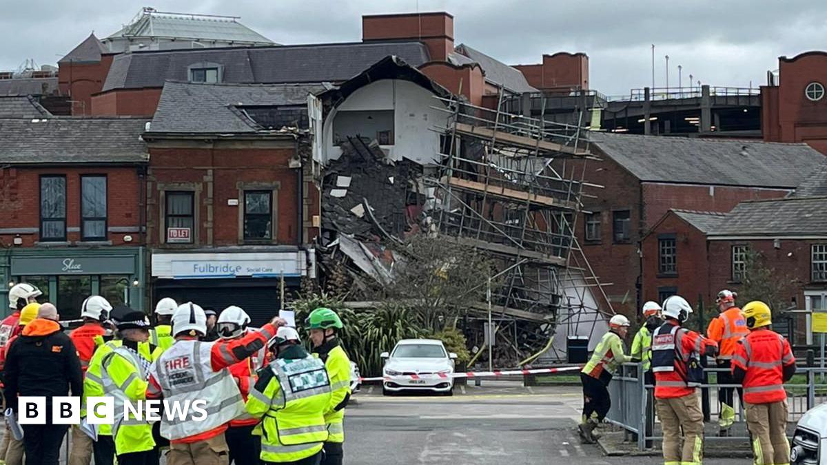 A partially collapsed building that shows roof tiles strewn over a pile of debris a scaffold hangs nearby. A number of emergency workers stand nearby.
