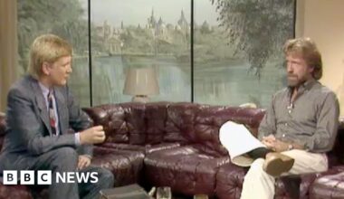 A man on the left wears a suit sitting on a couch facing a man on the right wearing a light brown shirt and light pants.