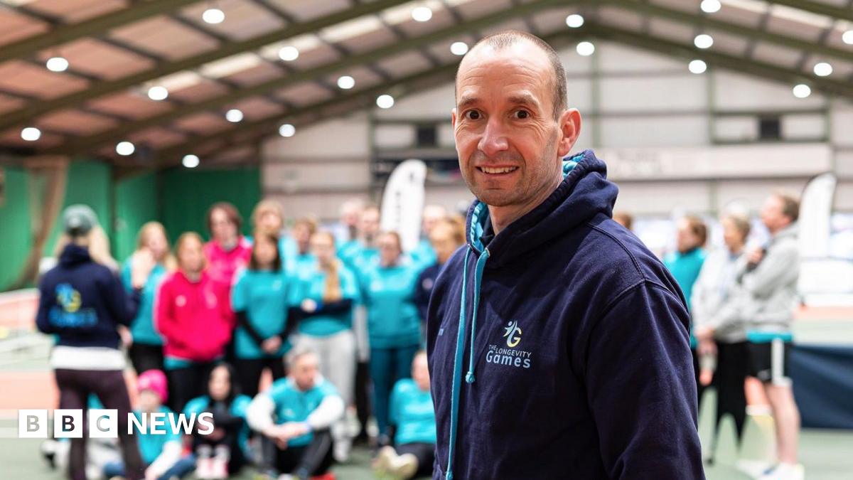 Tim Howe standing in front of a group of people in a sports hall. He smiles at the camera and wears a dark-blue hoodie which has The Longevity Games logo on it.