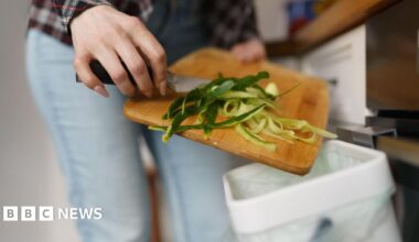Close-up of vegetable peelings being scrapped from a wooden chopping board into a small white food caddy lined with a white bag. A woman's hands can be seen holding the chopping board and knife being used.