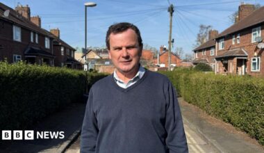 City of York Council's Labour deputy leader Pete Kilbane. He is looking straight at the camera, has receding brown hair, and is wearing a blue jumper and an open neck shirt. He is standing on a hedge-lined street in York. The sun is out and blue sky is in the background.