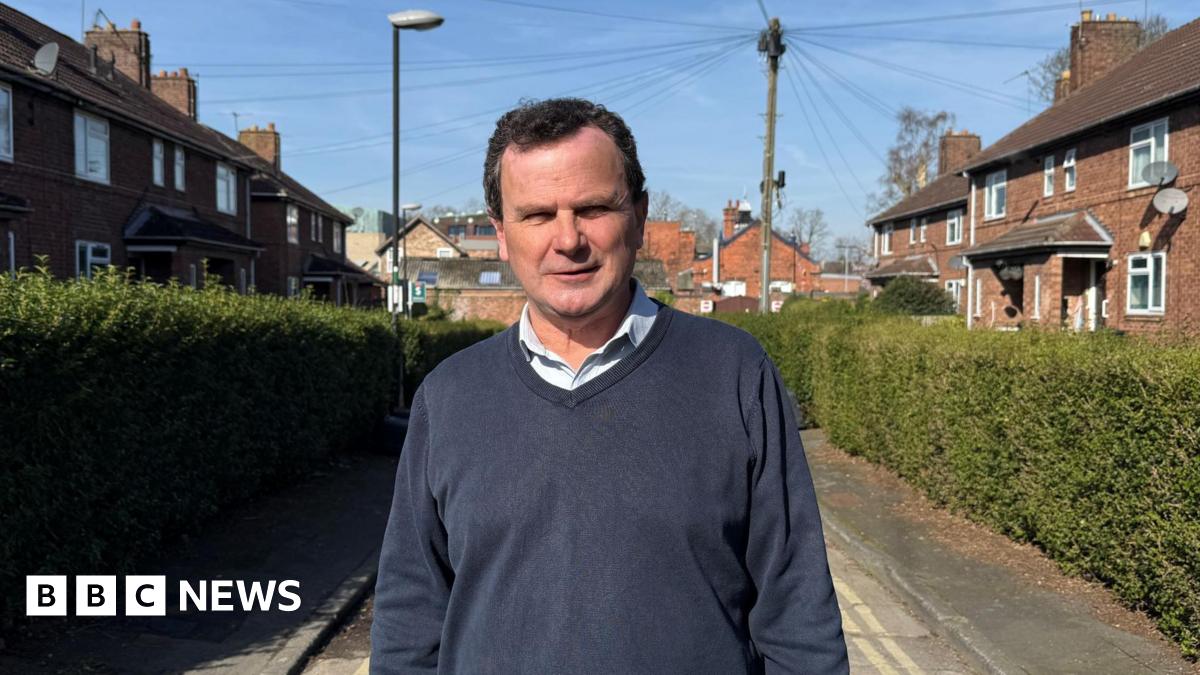 City of York Council's Labour deputy leader Pete Kilbane. He is looking straight at the camera, has receding brown hair, and is wearing a blue jumper and an open neck shirt. He is standing on a hedge-lined street in York. The sun is out and blue sky is in the background.
