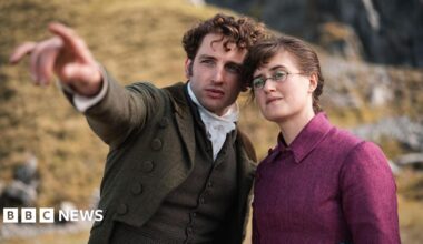Laurie Davidson and Ella Brucolleri in a still from The Other Bennet sister. Laurie is a man with brunette curly hair, a dark green jacket, a brown waistcoat and a white cravat. he is pointing to the left. Ella is wearing a cropped pink jacket and a blue tartan skirt.