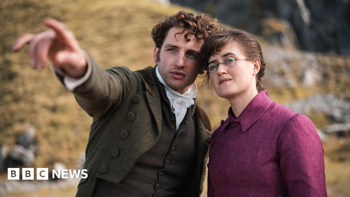 Laurie Davidson and Ella Brucolleri in a still from The Other Bennet sister. Laurie is a man with brunette curly hair, a dark green jacket, a brown waistcoat and a white cravat. he is pointing to the left. Ella is wearing a cropped pink jacket and a blue tartan skirt.