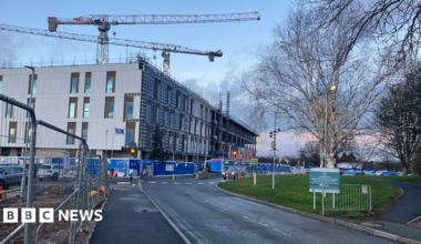 Building work at Royal Shrewsbury Hospital. Two cranes tower over a four-storey building, with metal fences on the left and a car park on the right