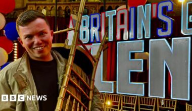 Sonny Green smiling at the camera holding a golden chair over his shoulder with the Britain's Got Talent logo and words in the background. He is wearing a khaki or golden-coloured jacket. He has short brown hair.