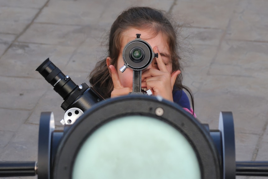 A young girl looks through a telescope at the sky.